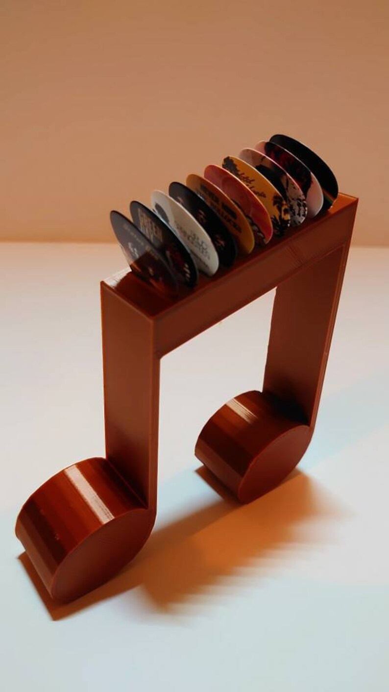 Puede incluir: Un soporte para p&uacute;as de guitarra de pl&aacute;stico marr&oacute;n en forma de nota musical con una fila de p&uacute;as de colores en la parte superior.