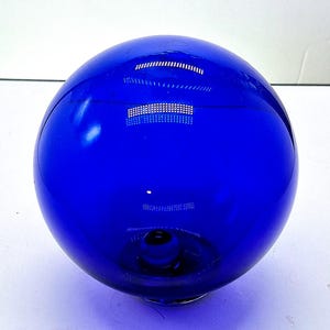 May include: A vibrant, cobalt blue glass sphere. The spherical object has a glossy, reflective surface. The sphere has a small opening at the bottom. The object is likely a decorative item or part of a larger piece.