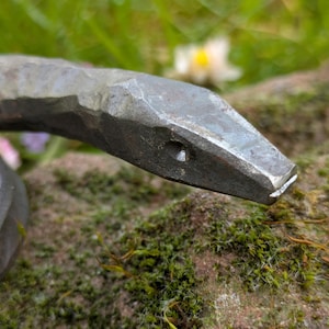 Op de afbeelding: Close-up van een handgesmede metalen slangenbeeld. De kop van de slang is naar beneden gericht, met een getextureerd oppervlak en een klein oogdetail. De sculptuur rust op een rots met mos, met een vage groene achtergrond.