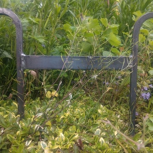 Może przedstawiać: Ciemnoszary, kutego żelaza, podpora do roślin ogrodowych. Podpora ma prostokątny, poziomy pręt z dwoma zakrzywionymi, haczykowatymi przedłużeniami. Podpora jest ustawiona w ogrodzie z zielonymi liśćmi.