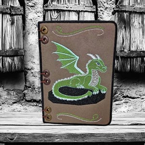 Peut inclure: Un journal marron avec une broderie de dragon vert. Le dragon a des ailes vertes et est en position couchée. Le journal est posé sur une étagère en bois devant un mur en pierre avec trois décorations en forme de tonneau en bois.