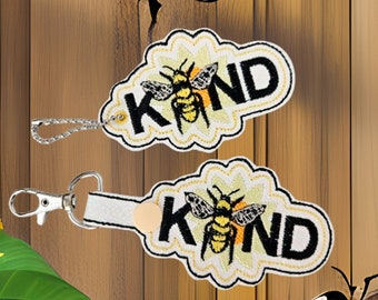Op de afbeelding: Twee gele en zwarte bij-thema sleutelhangers met het woord "KIND" op elk. De sleutelhangers bevinden zich op een houten achtergrond met zonnebloemen.