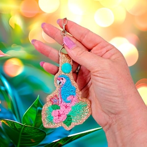May include: A pink flamingo keychain with blue and green embroidery details. The keychain is made of glitter fabric and has a turquoise blue bead at the top.