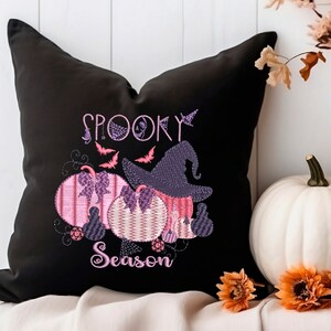 May include: Black throw pillow with embroidered pink and purple pumpkins, a witch's hat, and the text "Spooky Season". The pillow is sitting on a white surface with fall-themed decorations.