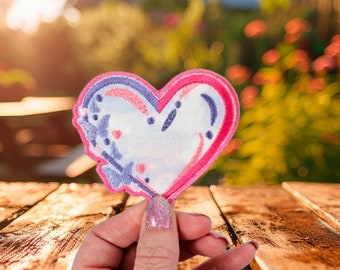 May include: A pink and blue heart-shaped patch with floral details. The patch is being held by a hand against a wooden background.