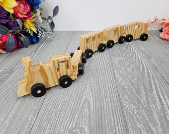 May include: A wooden toy train with four cars, including an engine, is made of natural wood with black wheels. The train is on a light gray wood surface.