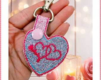 Puede incluir: Un llavero en forma de corazón de purpurina rosa y azul con un cierre de metal plateado y un botón rosa. El corazón tiene un diseño bordado rosa de dos corazones entrelazados con una flecha.