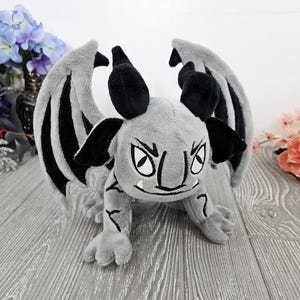 May include: A gray and black plush dragon with large wings, black horns, and white eyes. The dragon has a grumpy expression.