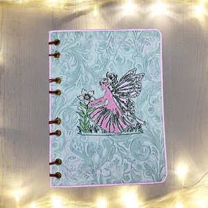 Op de afbeelding: Een lichtblauw notitieboek met een roze rand en een feeënontwerp. De kaft heeft een reliëf bloemenpatroon en een gestikte fee met een bloem. Het notitieboek is gebonden met koperkleurige ringen.
