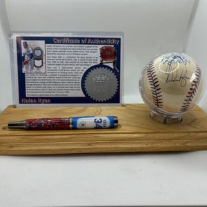 May include: A wooden display shelf holds a baseball in a clear case, a pen, and a certificate of authenticity. The baseball is white with red stitching and a signature. The pen has a red, white, and blue design. The certificate is in a clear frame.