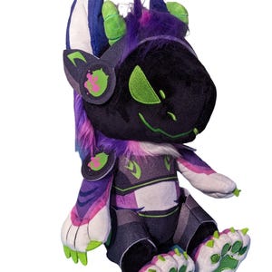 May include: A plush toy featuring a black, purple, and white creature with neon green accents. The character has large ears, a dark visor-like face with green eyes, and paw-shaped feet with green pads. It wears a headset and chest armor.