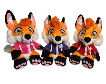 Op de afbeelding: Drie pluche vossen speelgoed met oranje en witte vacht, zwarte poten en grote, expressieve ogen. Elke vos draagt een hoodie in een andere kleur: roze, paars en rood. Het speelgoed is ontworpen met een cartoonachtige esthetiek.