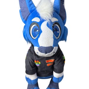 Może przedstawiać: Pluszowy niebiesko-biały wilk z białym kosmykiem futra i dużymi niebieskimi oczami. Wilk ma na sobie czarną kurtkę z kolorowymi naszywkami i napisem "WOLF OUT".