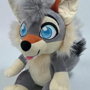 Op de afbeelding: Een pluche wolf met grote blauwe ogen, een roze tong en een pluizige grijze manen. De wolf heeft grijze en crèmekleurige vacht, met oranje accenten op de oren. Hij zit rechtop, met zijn poten en voeten zichtbaar.
