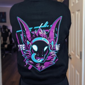 May include: Black sweatshirt featuring a purple and blue graphic of a furry creature with large ears and a glowing blue mouth. The text "VYTE ME" is printed on the sweatshirt.