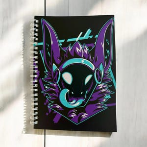 May include: A spiral-bound notebook with a black cover. The cover features a futuristic creature design in purple, teal, and white. The design includes a head with large ears and a protruding tongue against a black background.