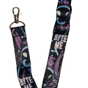 Pode incluir: Um cordão preto com um design roxo e azul que apresenta uma criatura de desenho animado com o texto "BYTE ME" repetido no cordão.
