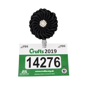 Puede incluir: Una roseta negra con un alfiler plateado y un centro de pedrería blanca está unida a una tarjeta verde y blanca con el texto "Crufts 2019" y el número "14276".