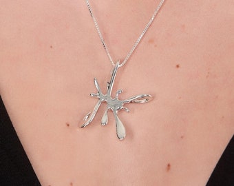 May include: A silver necklace with a large, abstract pendant shaped like a splash of liquid. The pendant is made of smooth, polished metal and has a modern, minimalist design.