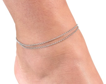May include: A silver double chain anklet. The anklet is made of two thin chains that are linked together. The anklet is worn on a person's ankle.