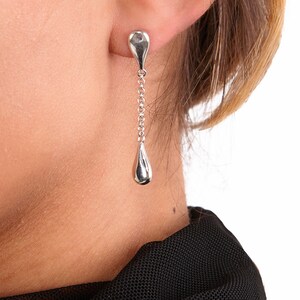 May include: Silver teardrop earrings with a delicate chain hanging from the top teardrop. The earrings are shown on a person's ear.
