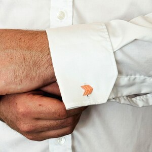 May include: Close-up of a white dress shirt with a cufflink. The cufflink is orange and shaped like an arrow. The shirt is buttoned and the sleeves are folded over the arms.