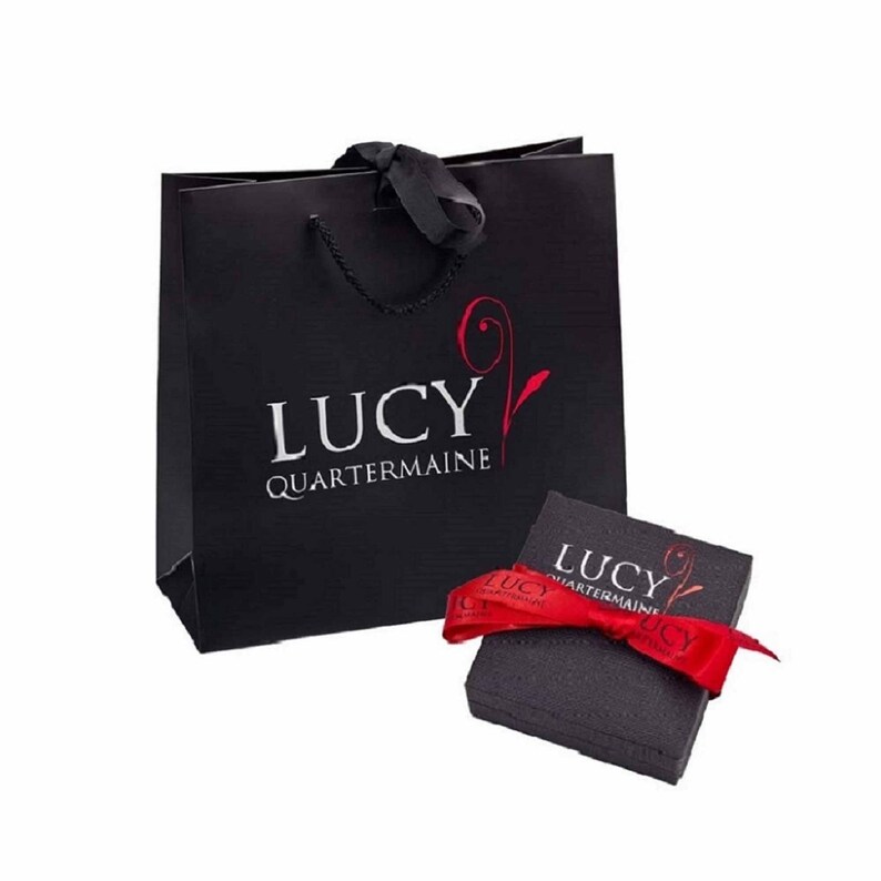 May include: A black gift bag with a red ribbon and the text "LUCY QUARTERMAINE" printed on the front. The bag is next to a black gift box with a red ribbon and the same text printed on the top.