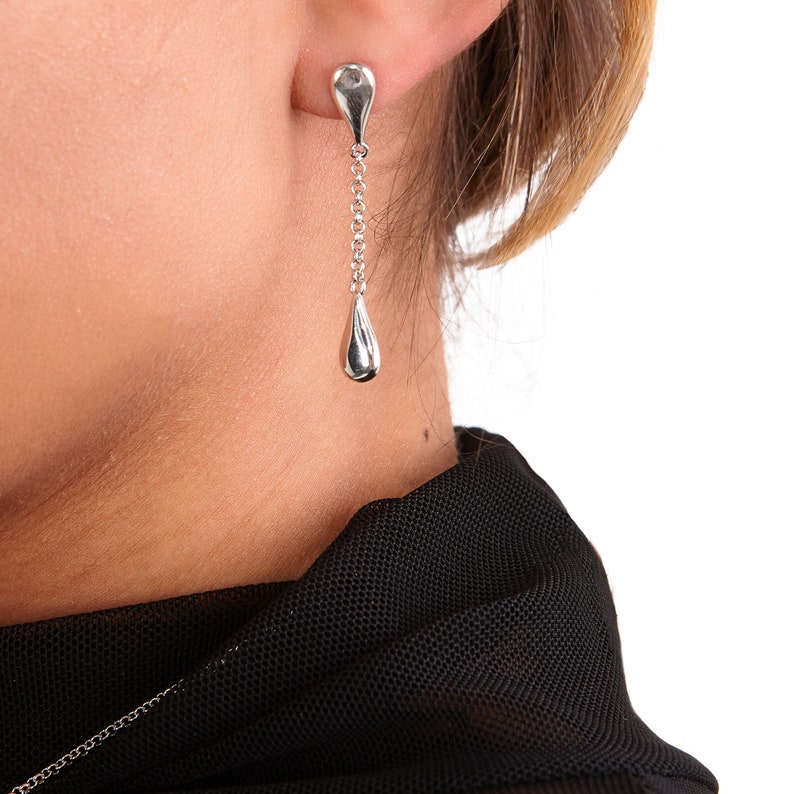 May include: Silver teardrop earrings with a delicate chain hanging from the top teardrop. The earrings are shown on a person's ear.