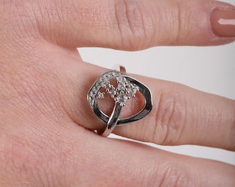 May include: A silver-toned ring featuring an open, asymmetrical oval frame. The band is adorned with a cluster of small, clear, round-cut stones set along one side of the oval, creating a sparkling accent against the polished metallic surface.