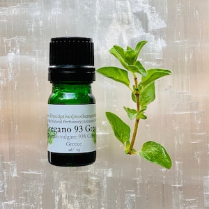 May include: A green glass essential oil bottle with a black cap, labeled "Oregano 93 Green." A sprig of fresh oregano with green leaves is next to the bottle. The background is a white, textured surface.