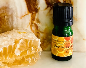 Può includere: Una bottiglia di vetro verde di olio essenziale "Beeswax Absolute" con tappo nero, accanto a un pezzo di favo. Lo sfondo è una superficie marmorizzata bianca e marrone.