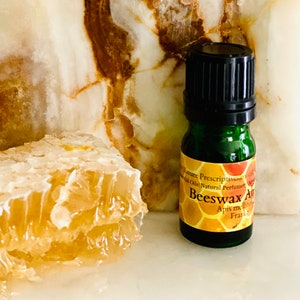 May include: A green glass bottle of "Beeswax Absolute" essential oil with a black cap, next to a piece of honeycomb. The background is a white and brown marbled surface.