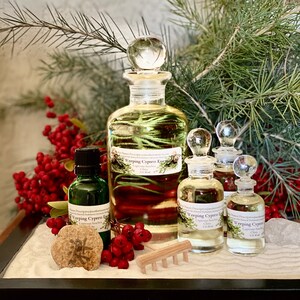 Puede incluir: Una colección de botellas de vidrio llenas de un líquido transparente, etiquetadas como "Weeping Cypress Essentials". Las botellas están dispuestas con bayas rojas, vegetación y un pequeño rastrillo de madera sobre un lecho de arena. El fondo es una pared clara y texturizada.