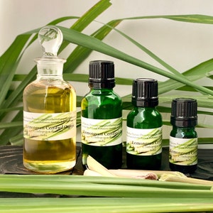 May include: A collection of essential oil bottles, including a large glass bottle with a stopper and three smaller green bottles with black caps. The bottles are labeled "Lemongrass Certified Organic" and are surrounded by lemongrass stalks and leaves.