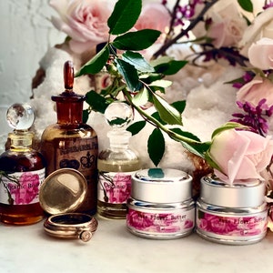 May include: An arrangement of apothecary bottles, jars, and a pocket watch, with a backdrop of roses and greenery. The bottles contain liquids, and the jars are labeled "Rose Flower Butter." The scene is illuminated by natural light.