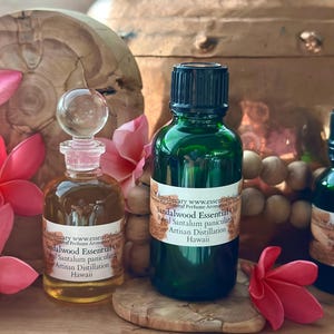 May include: Two glass bottles of sandalwood essential oil with labels, one clear with a glass stopper and one green with a black cap. The labels read "Sandalwood Essential Oil" and "Artisan Distillation Hawaii". Pink flowers and a wooden base are also visible.