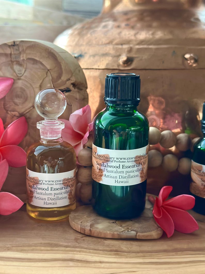 May include: Two glass bottles of sandalwood essential oil with labels, one clear with a glass stopper and one green with a black cap. The labels read "Sandalwood Essential Oil" and "Artisan Distillation Hawaii". Pink flowers and a wooden base are also visible.