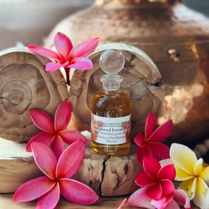 May include: A small glass bottle of sandalwood essential oil with a clear glass stopper, surrounded by pink and yellow plumeria flowers. The bottle sits on a wooden block, with a copper-colored vase in the background. The label reads "Sandalwood Essentials".