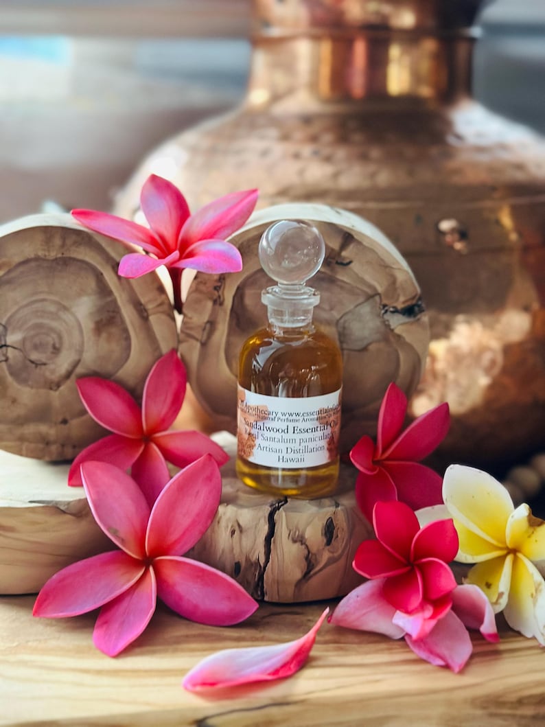 May include: A small glass bottle of sandalwood essential oil with a clear glass stopper, surrounded by pink and yellow plumeria flowers. The bottle sits on a wooden block, with a copper-colored vase in the background. The label reads "Sandalwood Essentials".