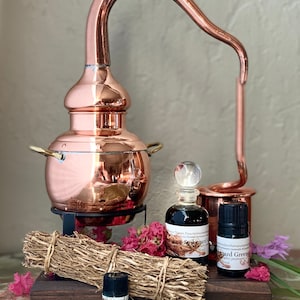 May include: A copper still with a pot, curved pipe, and small bottles. A bundle of dried herbs and pink flowers are arranged on a wooden base. The bottles contain essential oils, with labels that read "Spikenard".