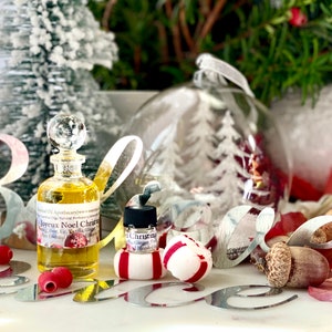 以下が含まれることがあります： クリスタルストッパー付きの透明なガラス香水瓶。黄色い液体が入っており、クリスマスの飾りの中に置かれています。「Joyeux Noel Christmas」と書かれたラベル。小さなボトル、ペパーミントキャンディー、装飾的なオーナメントも含まれています。