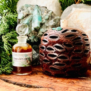 May include: A small glass bottle with a clear stopper and a brown liquid inside, labeled "Emerald Cypress Co." sits next to a decorative, dark brown, textured object. The background includes green foliage, white and green stones, all on a wooden surface.