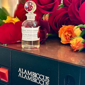 May include: A small glass bottle of Rose Otto Essential Oil with a clear stopper, set against a backdrop of red and orange roses. The bottle has a label with text. The base is black with the text "ALAMBICCUS ALAMBICCUS GAGGIA".