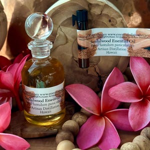 May include: A glass bottle of sandalwood essential oil with a clear glass stopper. The bottle is labeled with text. The image includes pink flowers, wooden beads, and a small vial of oil. The background is a wooden surface.