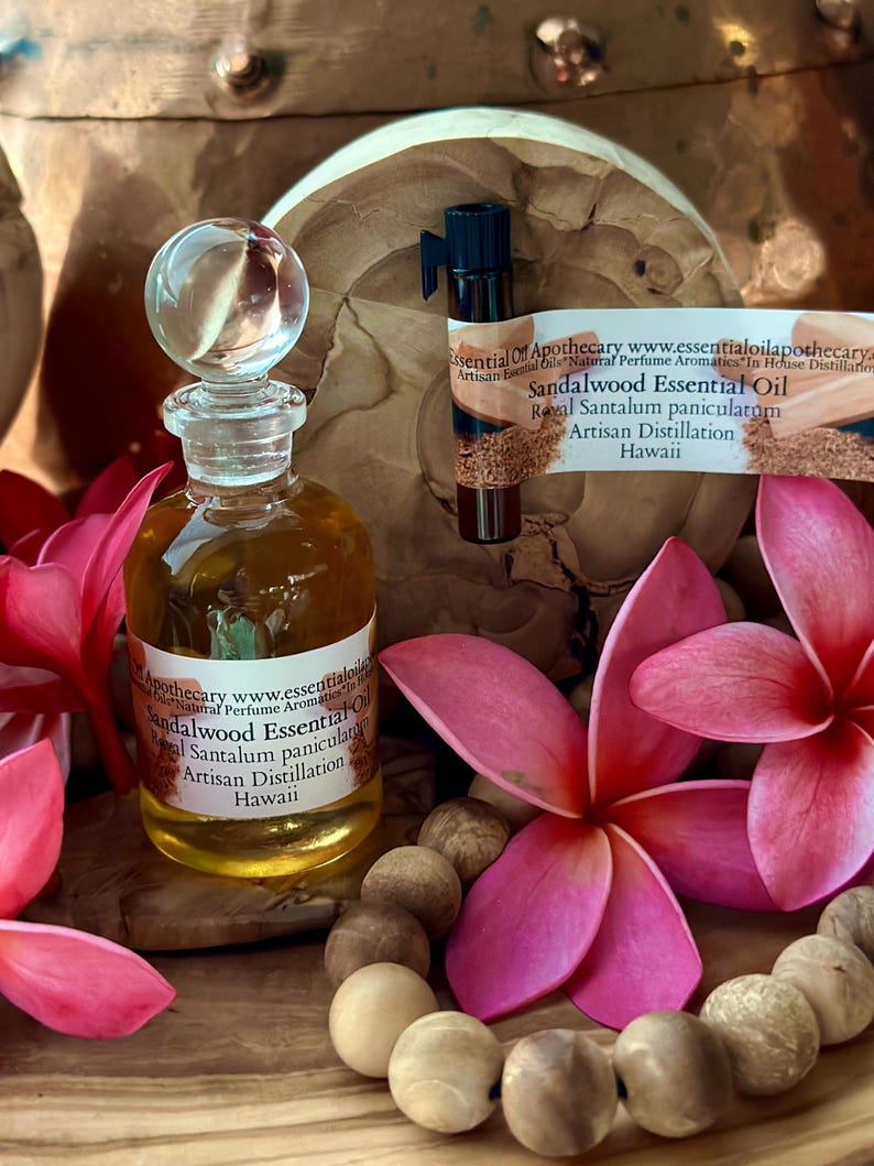 May include: A glass bottle of sandalwood essential oil with a clear glass stopper. The bottle is labeled with text. The image includes pink flowers, wooden beads, and a small vial of oil. The background is a wooden surface.