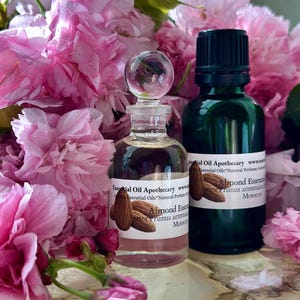 May include: Two essential oil bottles, one clear with a glass stopper and one dark green, are displayed amidst pink flowers. Labels read "Almond Essential Oil" and "Essential Oil Apothecary". The clear bottle has a label with almond illustrations.