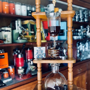 May include: A wooden cold brew coffee maker with a glass carafe and a clear top chamber. The coffee maker is filled with dark liquid, and coffee beans are scattered around the base. Small bottles of essential oils are also present.