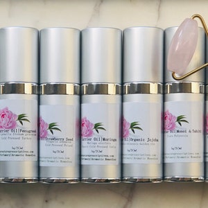 May include: An assortment of silver-topped bottles with labels featuring a pink peony design, alongside a rose quartz face roller with a gold handle. The bottles contain various carrier oils, each labeled with its botanical name.
