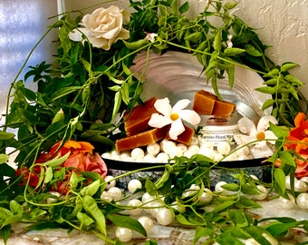 May include: A display of jasmine floral wax and soap bars, surrounded by white pearls, green vines, and colorful flowers. The wax is in a small jar and the soap is cut into squares. The arrangement is on a decorative tray.