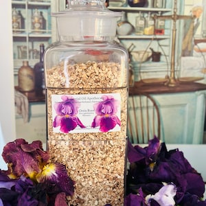 May include: A clear glass apothecary jar filled with beige, dried orris root. The jar has a glass stopper and a label with purple iris flowers. Purple iris flowers are arranged around the jar, with a vintage apothecary illustration in the background.